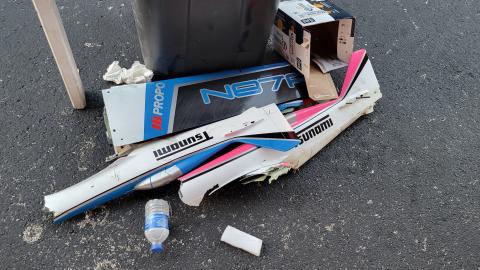 Broken model airplane pieces with colorful decals on asphalt near a trash bin.