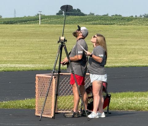 Two people watch the sky with a camera on a tripod in a grassy field.