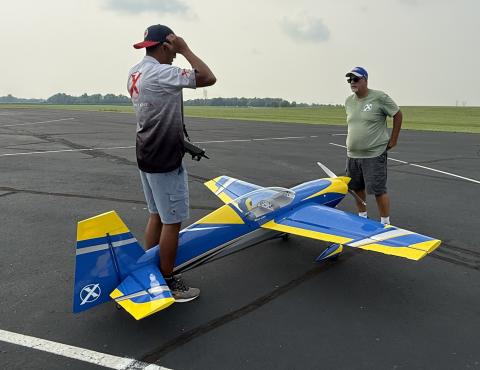 Two men standing by a large blue and yellow model plane on asphalt.