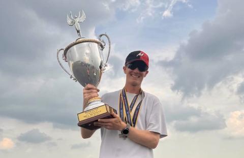 Man smiling, holding a large trophy outdoors, wearing a red and black cap.