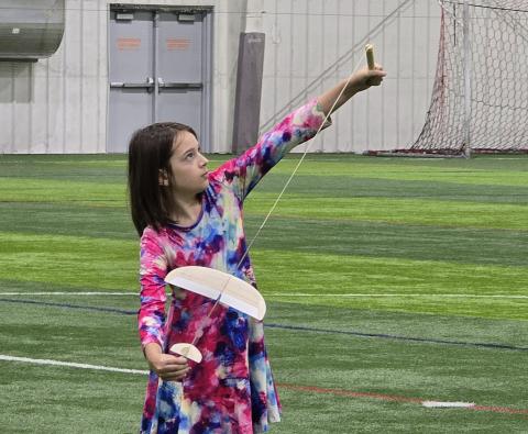 Young girl aiming a wooden bow and arrow indoors on a grass field.