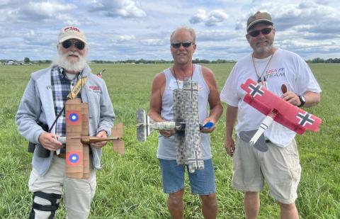 Three men holding remote-controlled biplanes in a grassy field.