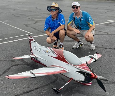 Two men crouching beside a large remote control airplane on a parking lot.