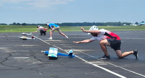 Three people launching model planes on a runway, wearing helmets.