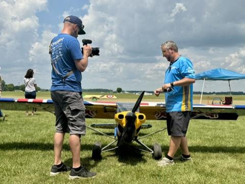 Man films another near small model plane on grass field.