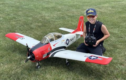 Young boy in cap kneels on grass beside a large model Navy aircraft, smiling and holding a remote.