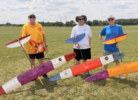Three men standing on grass with colorful model gliders.