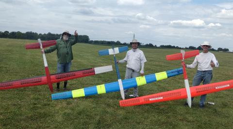 Three men holding colorful model gliders on grassy field.