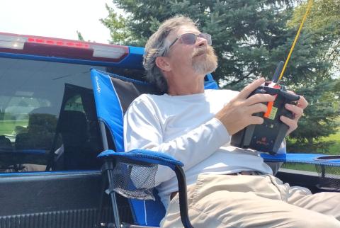 Man in sunglasses holding a remote control, sitting relaxed in a truck bed.