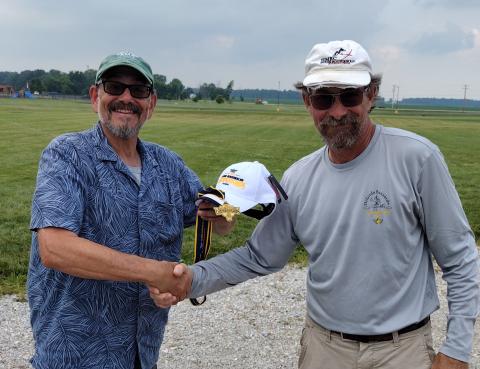 Two men smiling, shaking hands, one holding a medal outdoors.