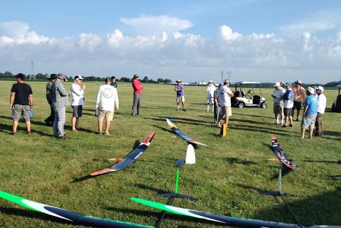 Model gliders on grass with a group of people in a field, talking and observing.