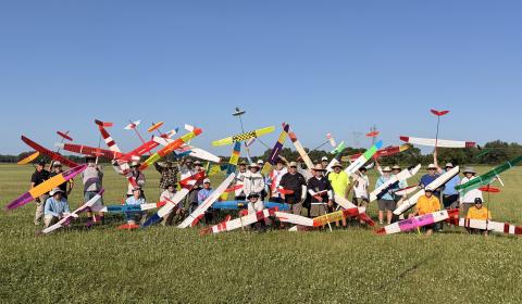 Group of people holding colorful model gliders in a grassy field.