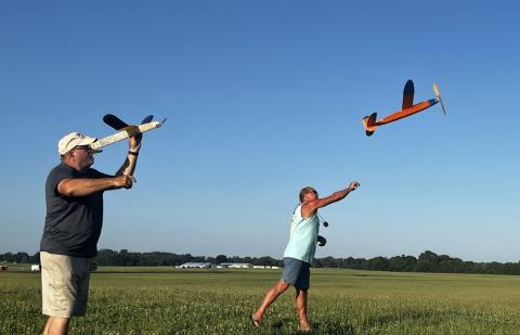 Two men launch model gliders in a grassy field under a clear blue sky.