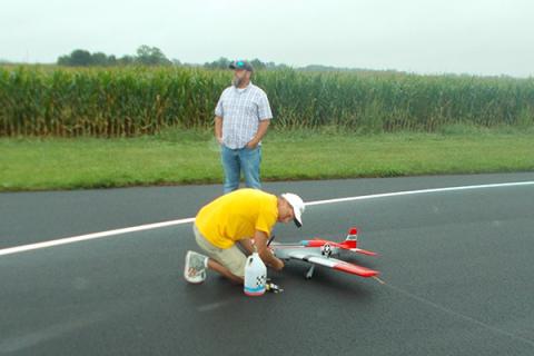 Man in yellow kneels to adjust model airplane on a runway; another man stands nearby.