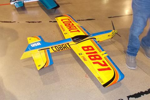 Yellow and blue model airplane on the floor near people’s feet.