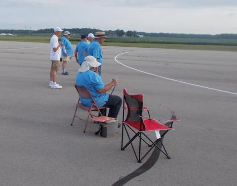 People in blue shirts, two seated, on an empty runway with overcast skies.