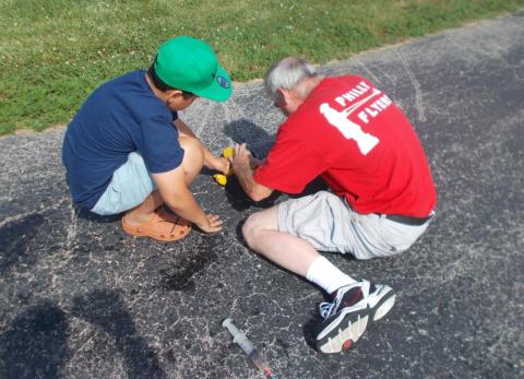Two people kneeling on pavement, fixing a black RC car.