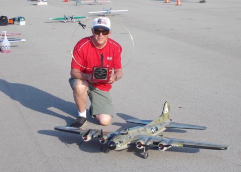 Man kneeling beside a model aircraft on pavement, holding a plaque, wearing sunglasses and a cap.