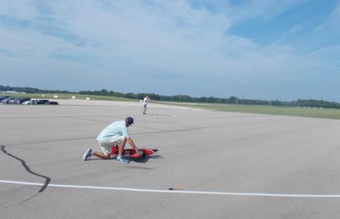 Person preparing model airplane on wide runway under a clear blue sky.