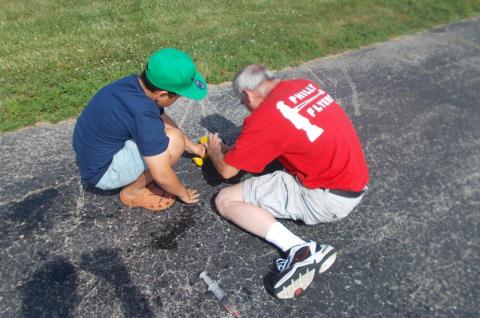 Two people fixing a toy car on a driveway in sunlight.