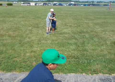 Two boys playing in a grassy field on a sunny day, with cars in the distance.