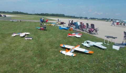 Model airplanes lined up on grass near a runway, with people sitting nearby.