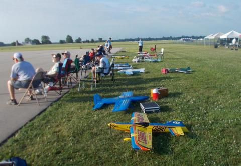 Model airplanes lined up on grass, people seated nearby on a runway edge.