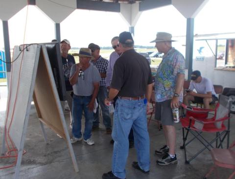 Group of men in casual attire gathered around a board in an open shelter.