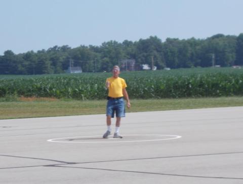 Man in yellow shirt stands on airfield, cornfield in background.