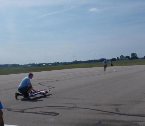 Man crouching near a model airplane on a runway, with a grassy field in the background.
