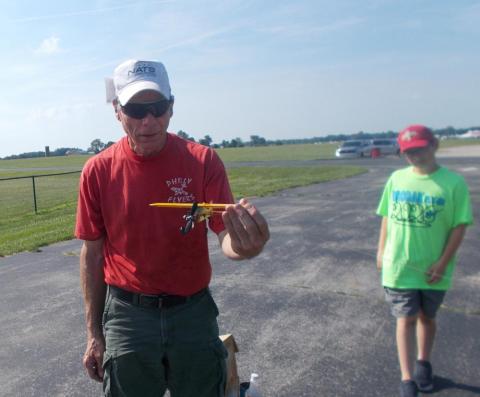 Man in red shirt holding a model plane, boy in green shirt walking nearby.
