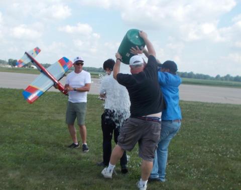 Three men pour ice over a man holding a toy plane on a grassy field.