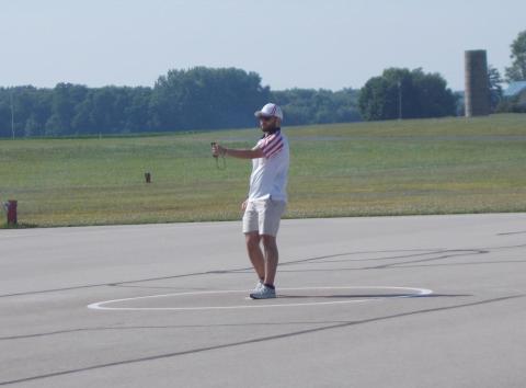 A man standing on a large outdoor paved open space with fields and trees in the background.
