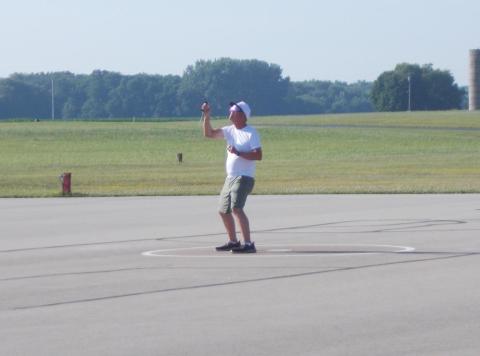 Man flying a kite on a large open area with grass and trees in the background.
