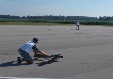 Man kneeling on an airstrip with a model plane, another person in the background.