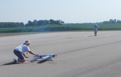 Man kneeling with a model plane on a runway, another person stands in the distance.