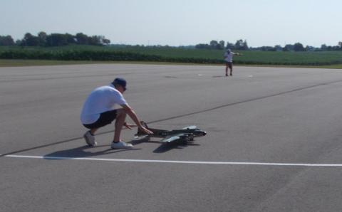 Man crouching on tarmac with a model airplane, another person in the background.