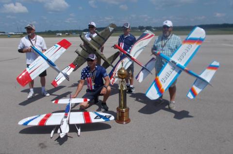 Five people with model airplanes on an airstrip, holding a large trophy.