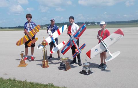 Four people holding model airplanes with trophies on the ground.