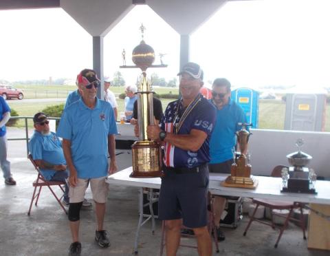 Man holding large trophy, surrounded by smiling people in a pavilion.