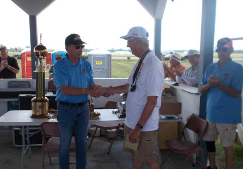 Two men shaking hands beside a large trophy under a pavilion.