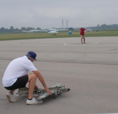 Man in shorts kneeling on runway with a model jet, another person standing in background.
