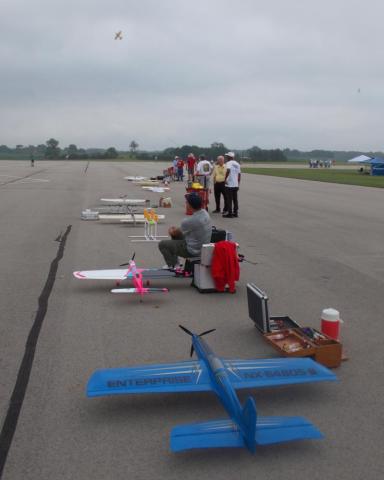 Model airplanes lined up on a runway with enthusiasts preparing for flight.