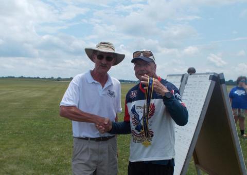 Two men outdoors, one holding medals, both smiling and shaking hands.