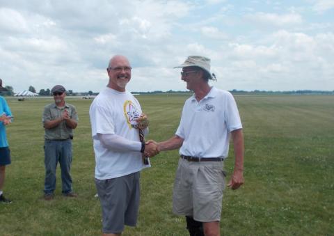 Two men smiling and shaking hands on a grassy field, with others clapping in the background.