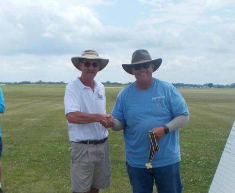 Two men in hats shake hands on a grassy field, holding a gold medal with ribbon.