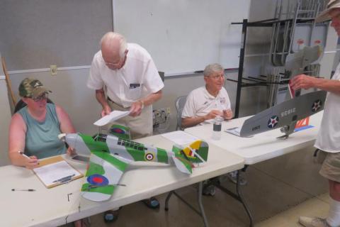 Model airplane contest with judges scoring entries on a table.