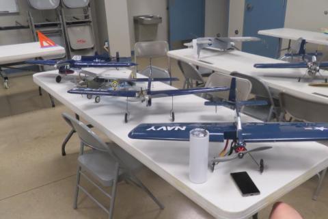 Model airplanes on tables in a workshop room with chairs and tools.