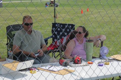 Two people sitting behind a table with electronics, outdoors behind a wire fence.
