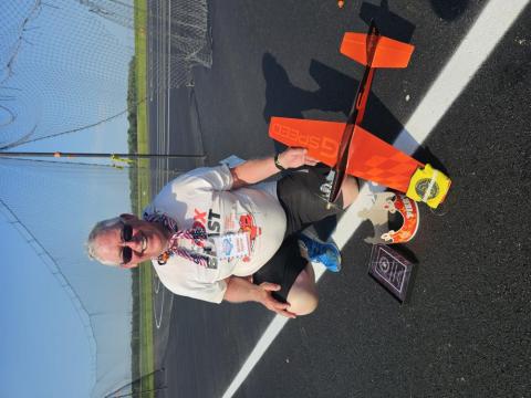 Man kneeling on a sunny asphalt track with a model airplane and a trophy.